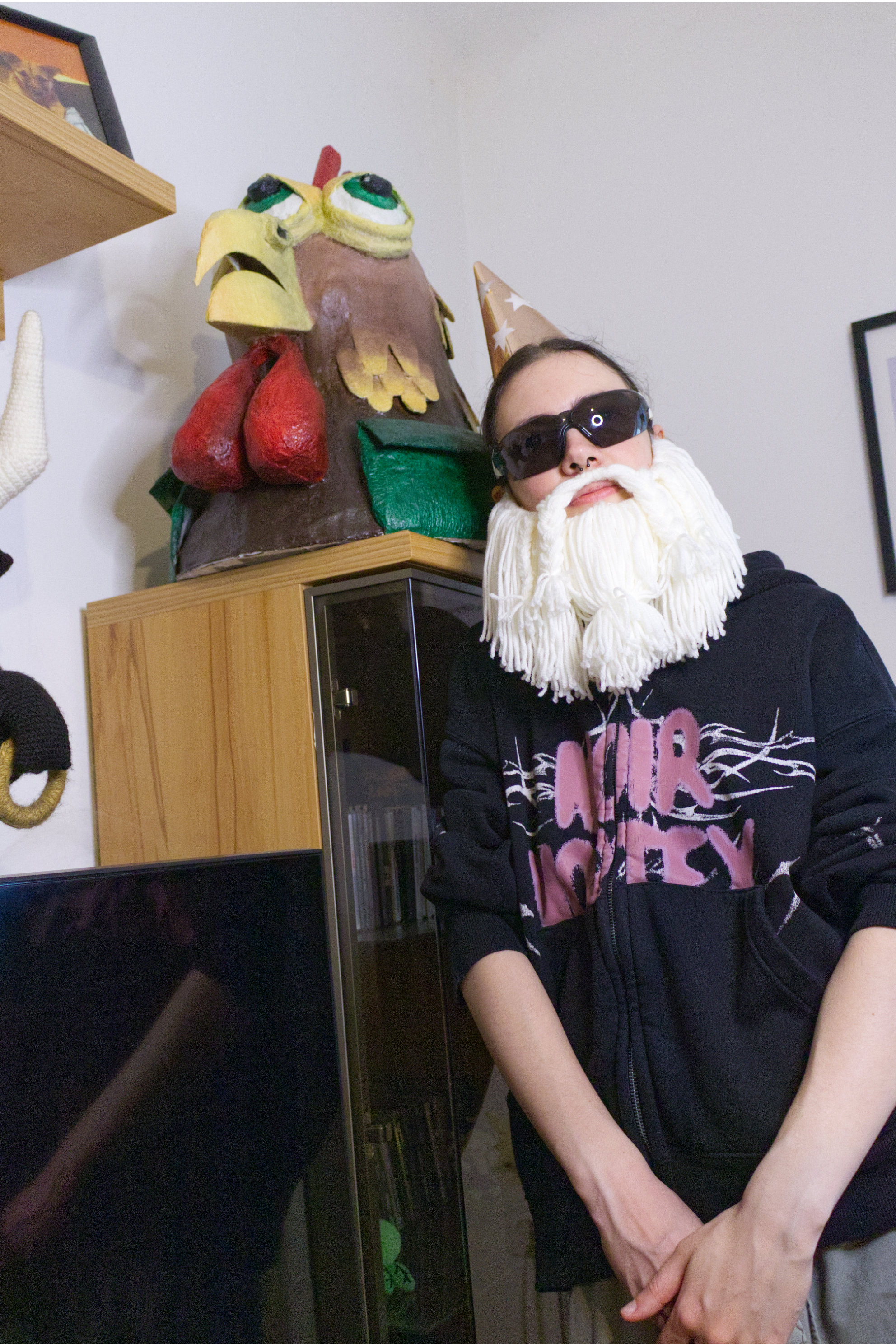 The Crafting Wizard wearing a beard, hat, and sunglasses stands in front of a colorful radioactive chicken sculpture in the background.
