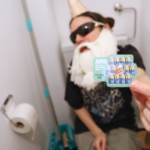 Cross-stitch ADHD “All Ducks Have Daddy Issues” sticker held in the Crafting Wizard’s hand while he sits on a toilet, shown in a funny staged scene with the Crafting Wizard as the main subject.