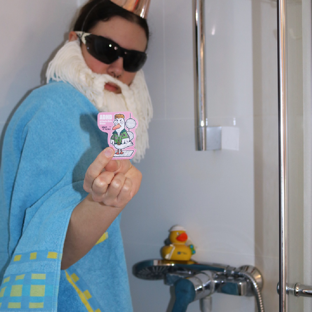 Sticker “ADHD - All Ducks Hate Discos” held by the Crafting Wizard standing in the shower wearing a towel, with a rubber duck in the background in a funny staged scene.