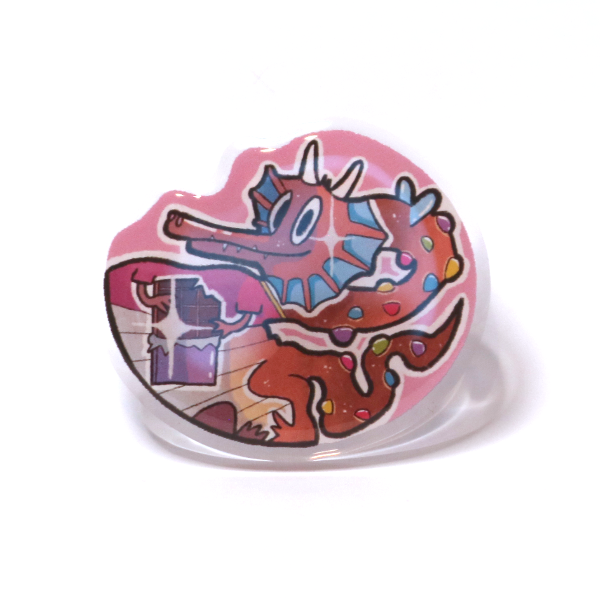 Acrylic pin of a cute chocolate dragon with a red T-shirt, holding chocolate and covered in colorful chocolate drops instead of horns and scales.