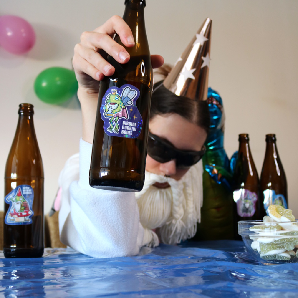 Drunk Fairy Frog Sticker placed on a bottle, shown in a funny party scene with the Crafting Wizard at a kids birthday table, decorated with a colorful tablecloth and balloons in the background.