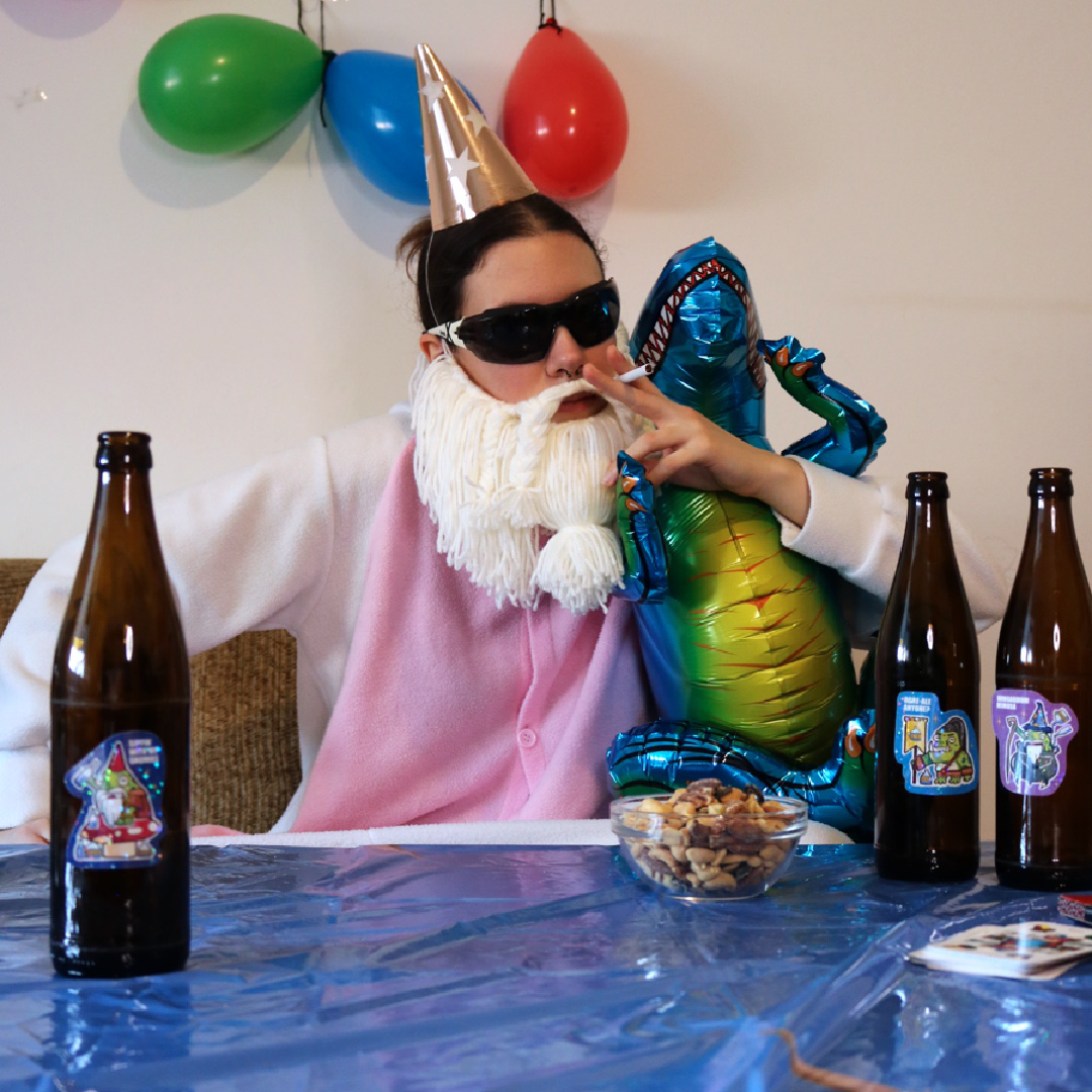 Drunk Gnome Frog Sticker placed on a bottle, shown in a funny party scene with the Crafting Wizard at a kids birthday table, decorated with a colorful tablecloth and balloons in the background.
