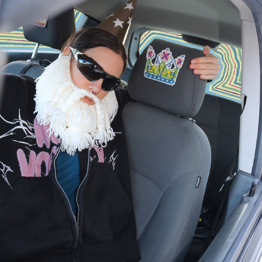 Cross-stitch Fairy Crown placed on a car seat neck support for a “passenger princess” setup, with the Crafting Wizard present, a humorous staged scene.