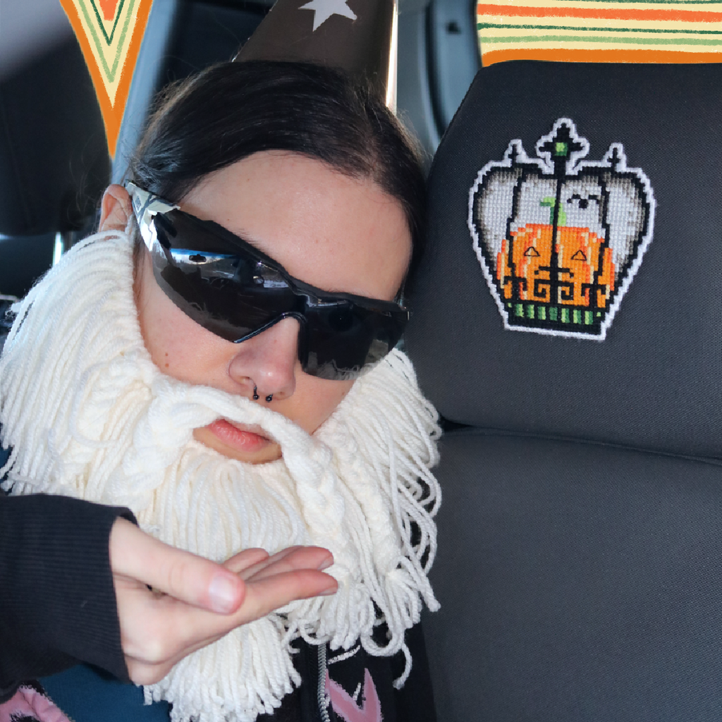 Cross-stitch Halloween Crown placed on a car seat neck support for a “passenger princess” setup, with the Crafting Wizard present, a humorous staged scene.