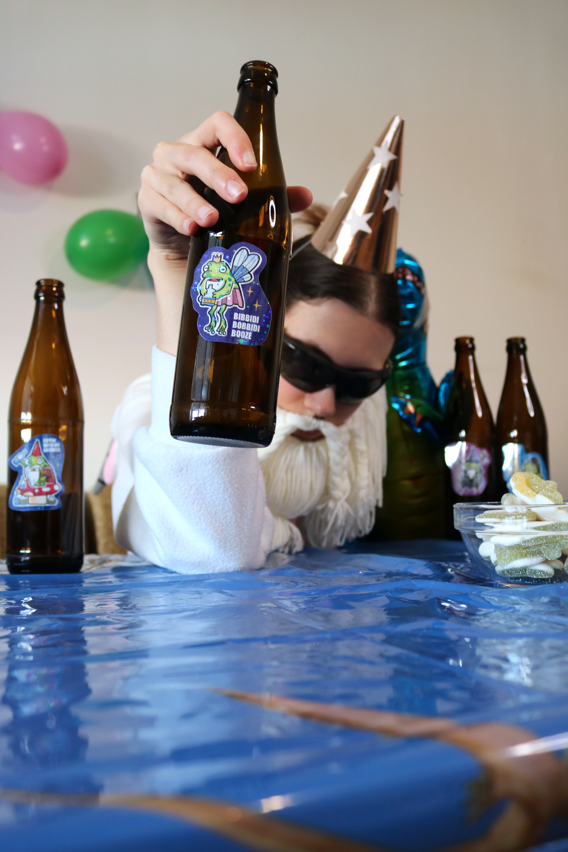 Drunk Fairy Frog Sticker placed on a bottle, shown in a funny party scene with the Crafting Wizard at a kids birthday table, decorated with a colorful tablecloth and balloons in the background.
