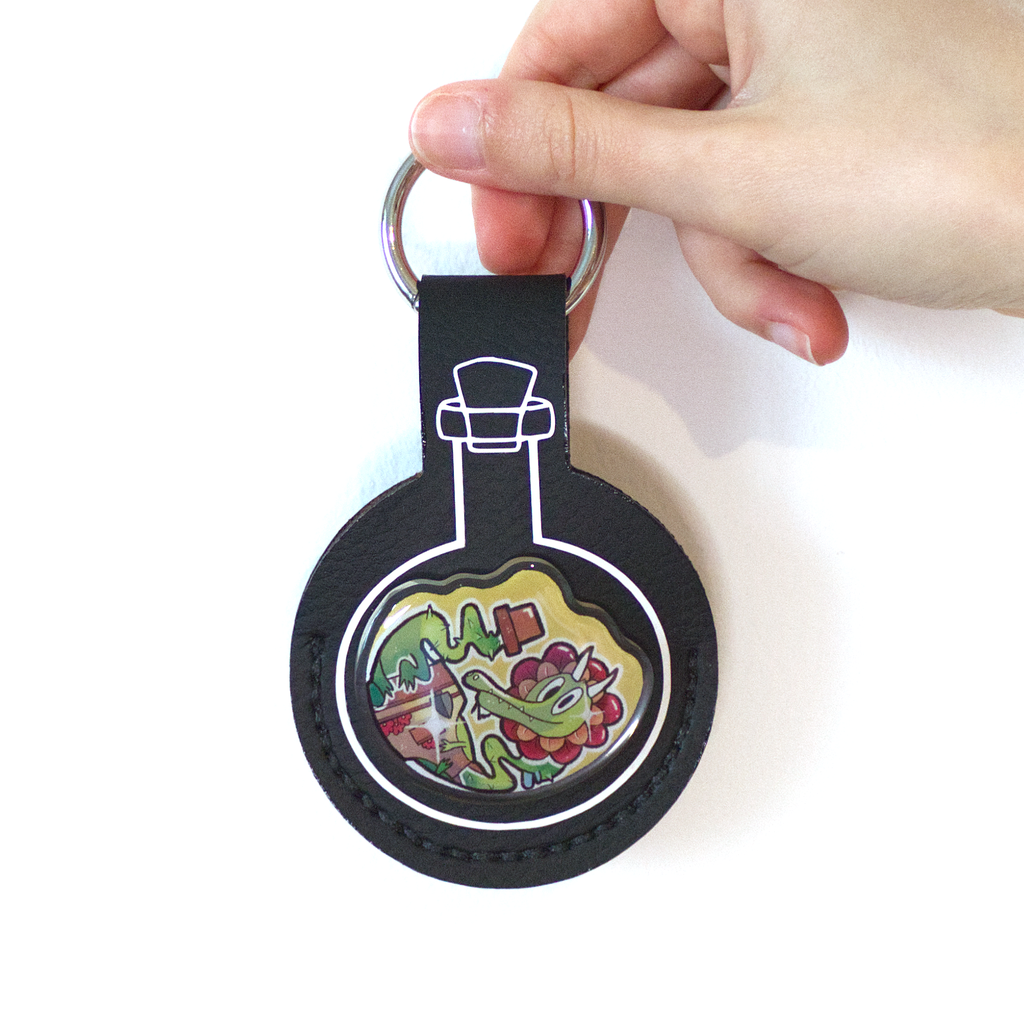 Black faux-leather potion keychain with a resin-finished acrylic pin of a plant dragon growing from a flower pot, wearing a gardening apron with tools.