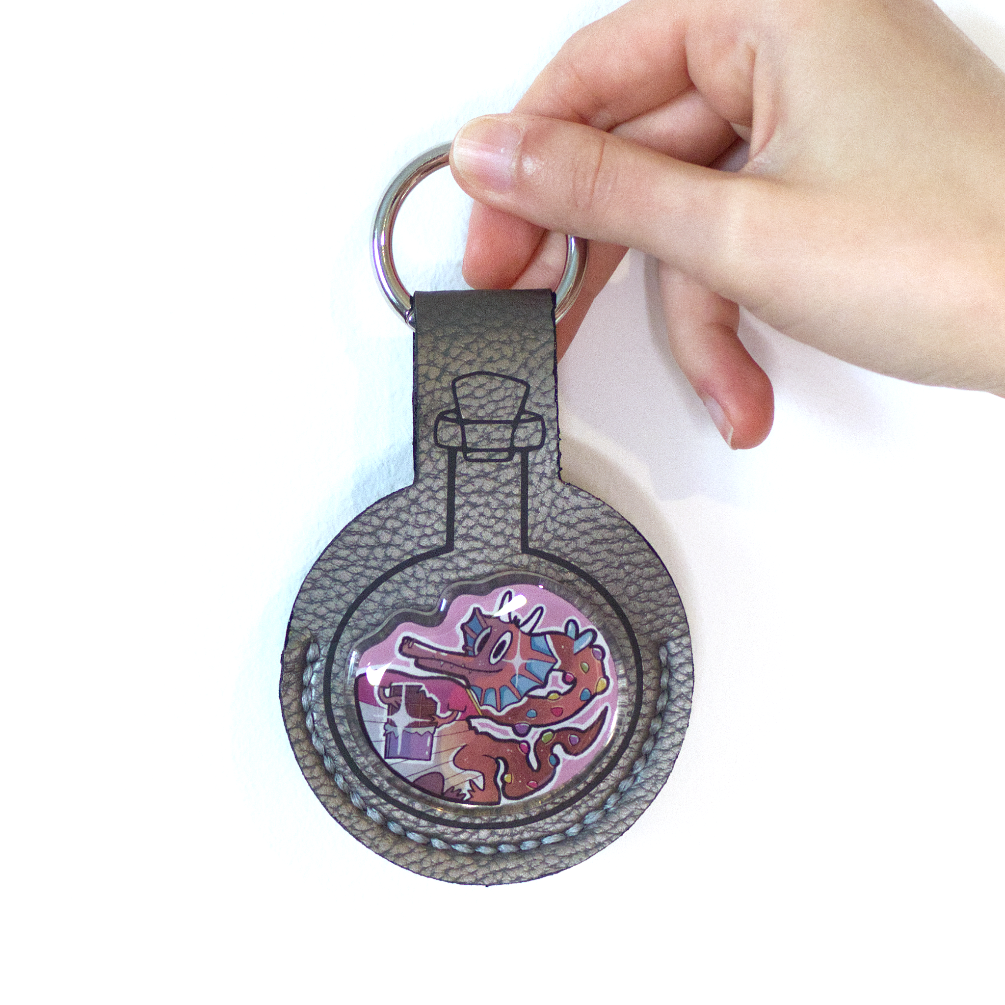 Grey faux-leather potion keychain with a resin-finished acrylic pin of a cute chocolate dragon wearing a red shirt and holding chocolate pieces.