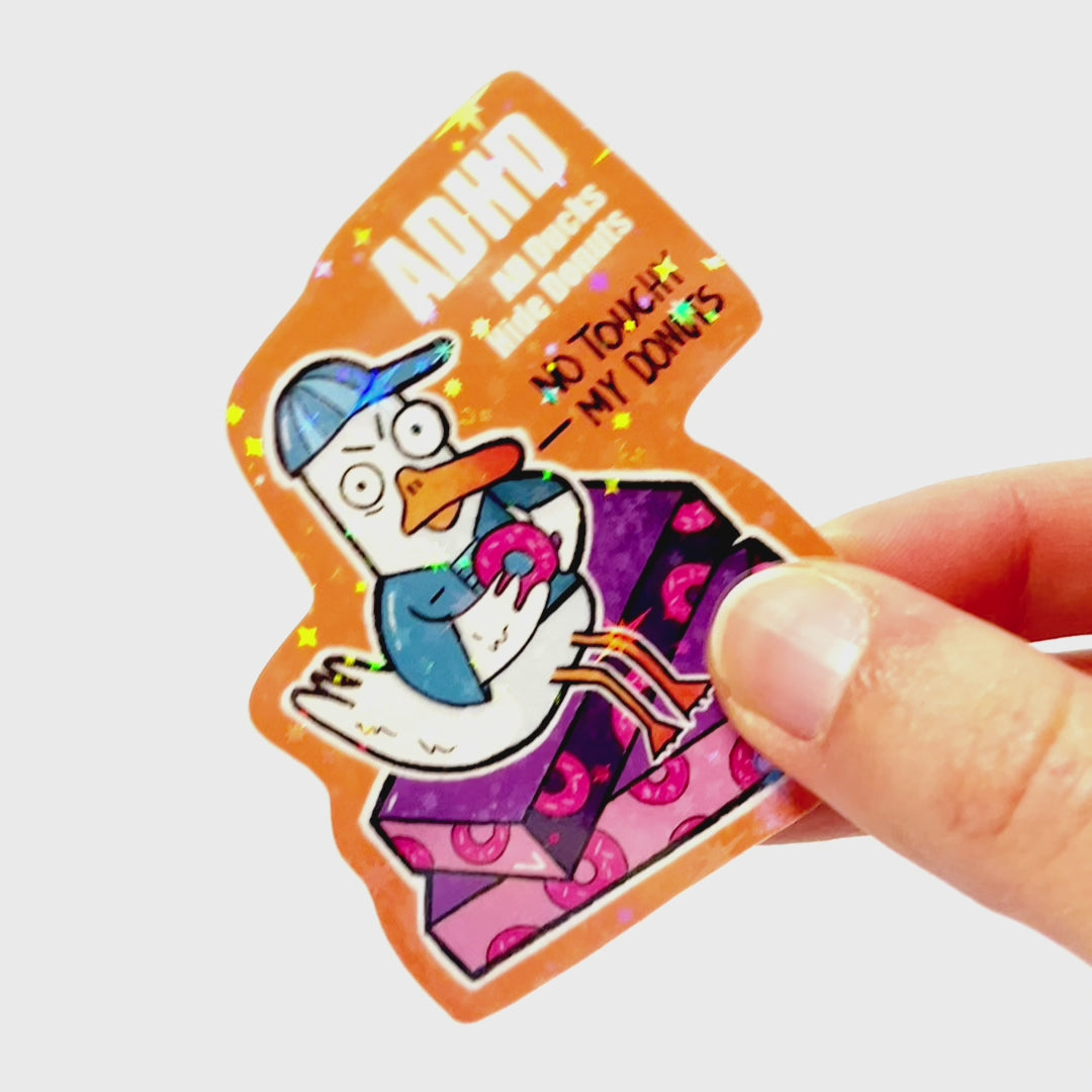 Video: Sticker of an angry duck in blue clothes sitting on donut boxes, holding a donut, with the slogan ‘ADHD – All Ducks Hide Donuts.’