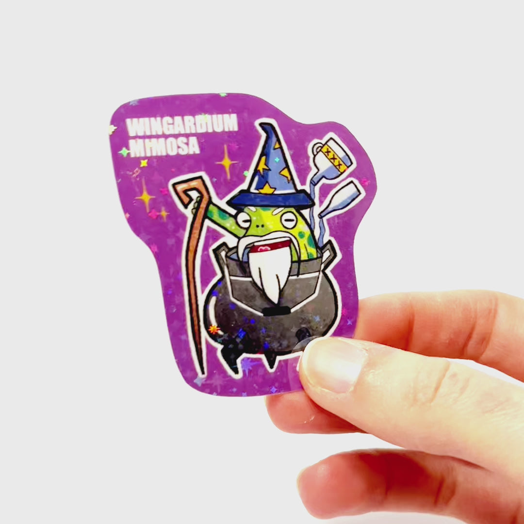 Video: Sticker of a drunk wizard frog with a beard and hat, sitting in a cauldron and magically pouring bottles into it, with the slogan ‘Wingardium Mimosa.’