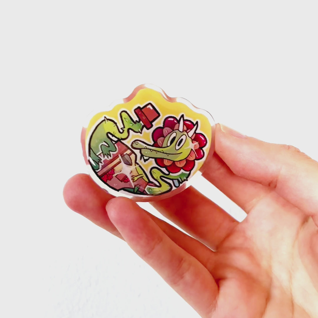Video: Acrylic pin of a plant dragon growing from a flower pot, wearing a gardening apron with tools, a flower around its head, and rose-like thorns.

