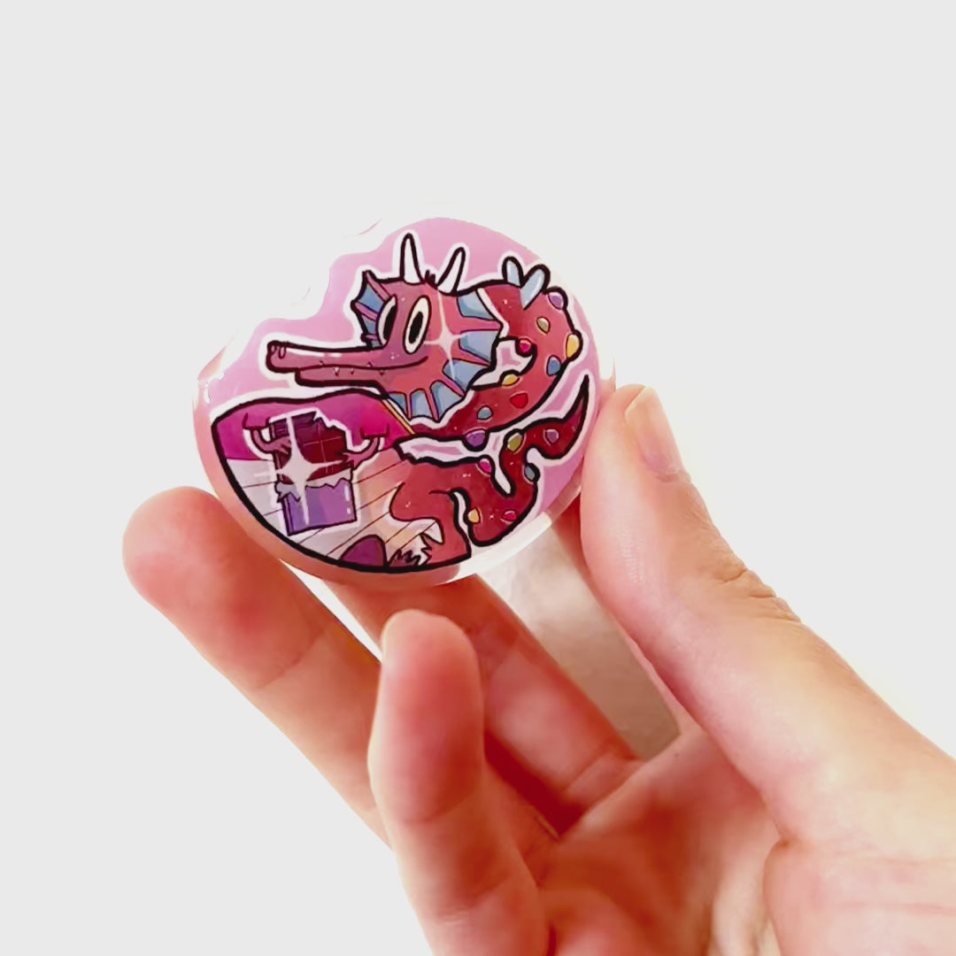 Video: Acrylic pin of a cute chocolate dragon with a red T-shirt, holding chocolate and covered in colorful chocolate drops instead of horns and scales.