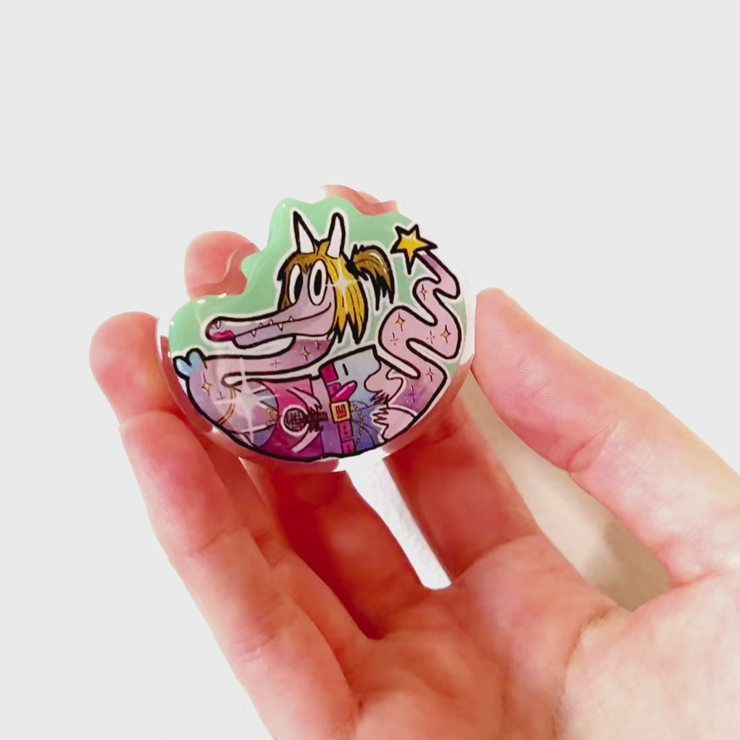 Video: Acrylic pin of a Y2K-style dragon with blonde bob hair, holding a microphone, wearing trendy Y2K fashion, with a star at the tip of its tail.